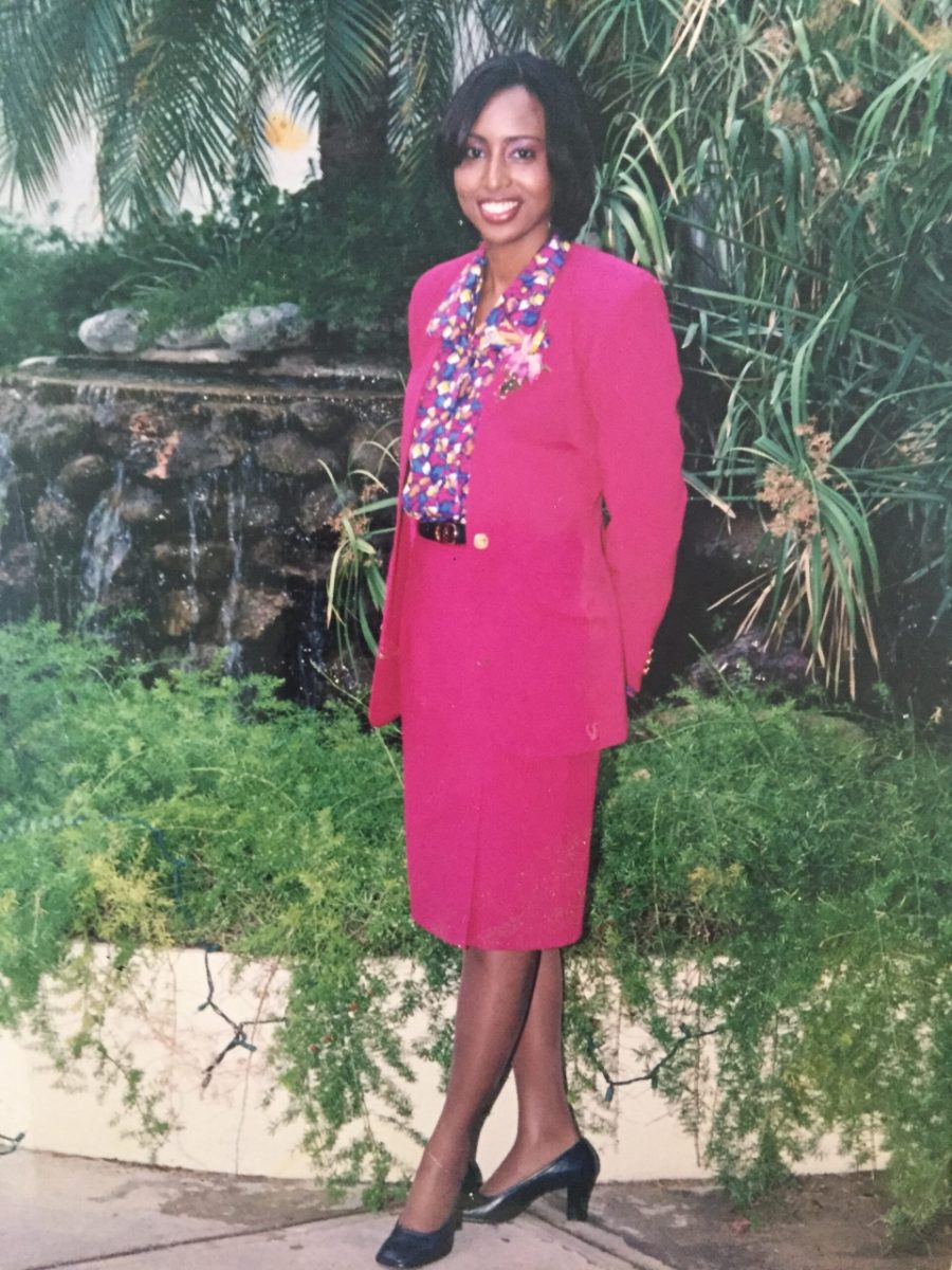 Yanique Grant as a young Air Jamaica flight attendant in a pink suit, standing in a tropical garden
