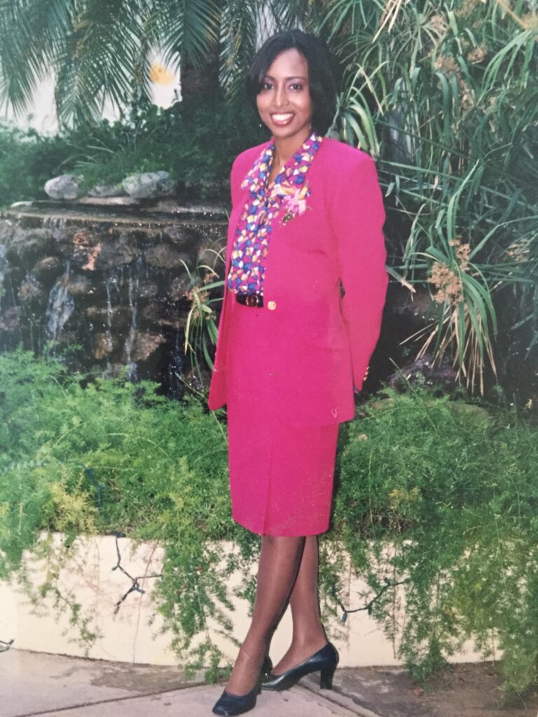 Yanique Grant as a young Air Jamaica flight attendant in a pink suit, standing in a tropical garden